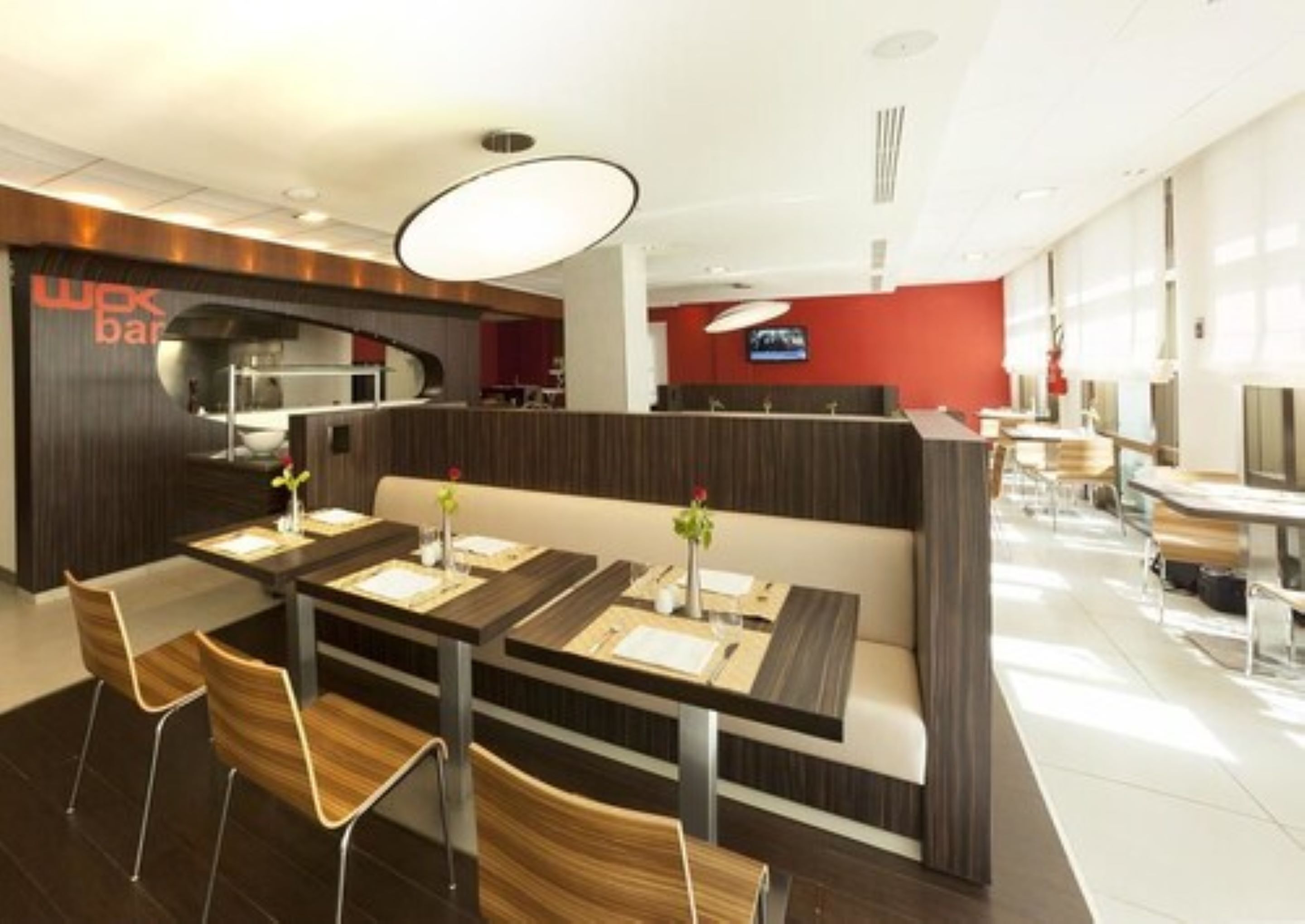 Un restaurante moderno con un comedor central que presenta mesas y sillas de madera, una zona de bar con acabados en madera oscura y una pared de acento roja.