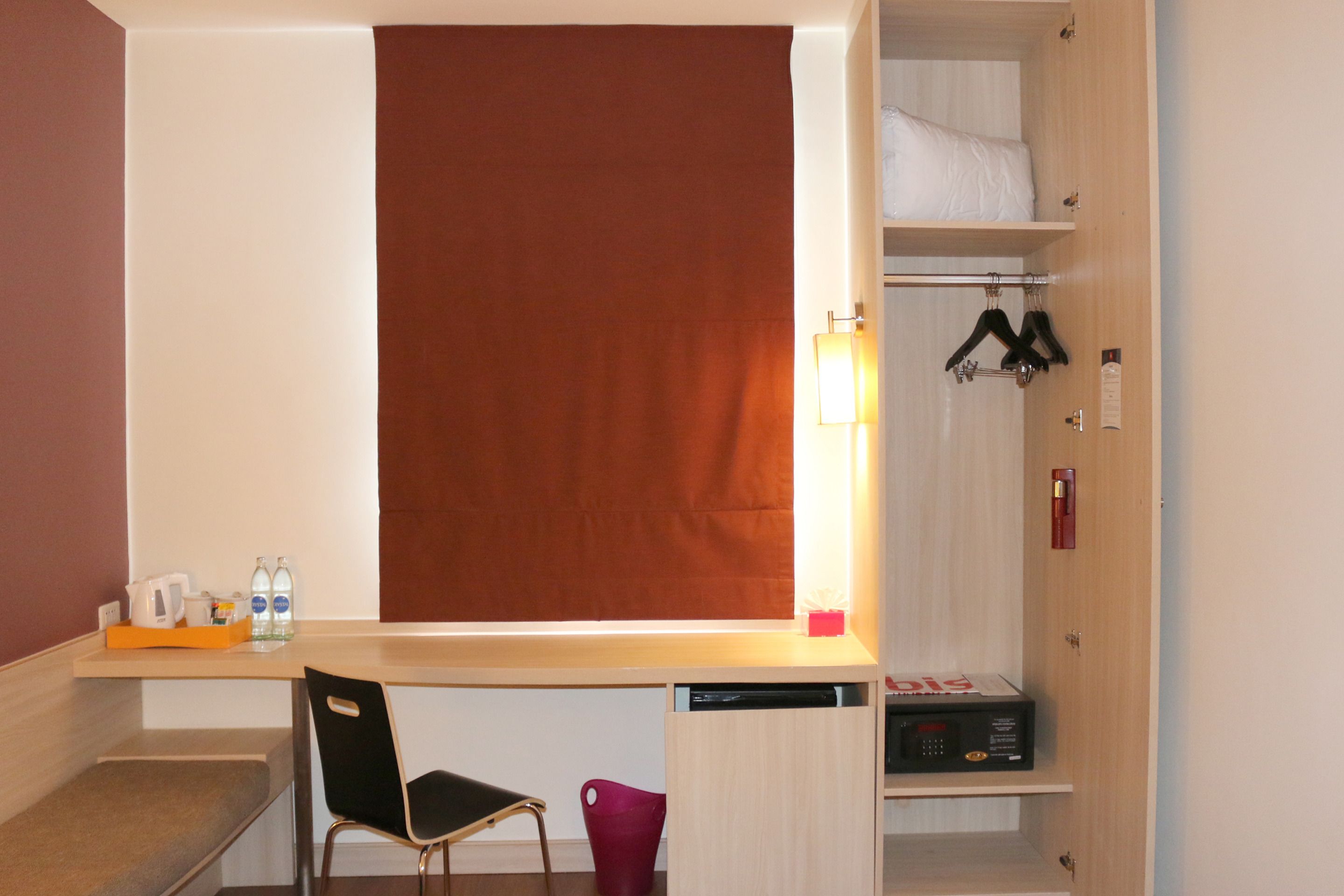 Hotellihuoneessa on puinen työpöytä, tuoli, pieni pöytä, jossa on vedenkeitin ja pulloja, punainen ämpäri, hylly, jossa on laatikko, ja vaatekaappi, jossa on henkareita.