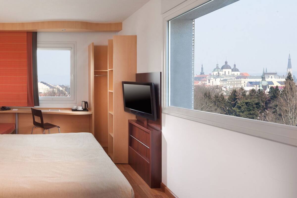 Una habitación de hotel con cama, escritorio, silla, televisión y ventana con vista a la ciudad.