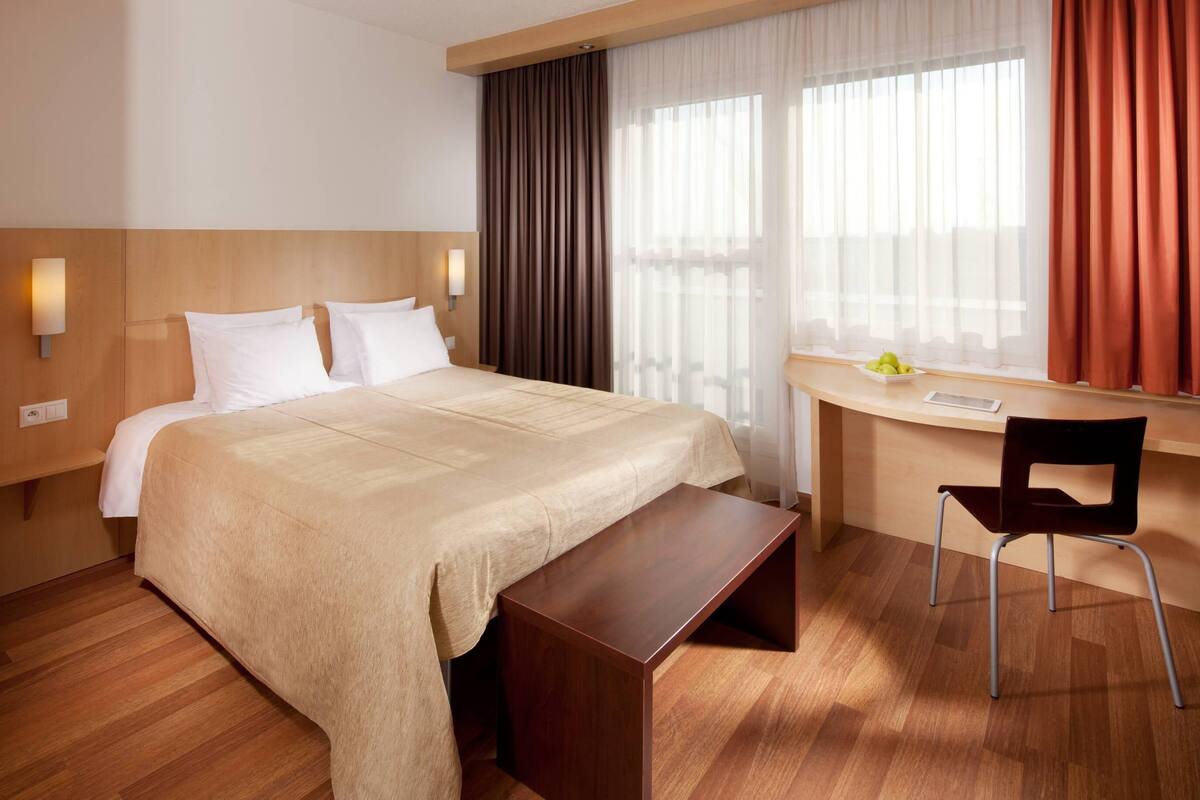 Habitación de hotel con cama, mesita de noche, silla, escritorio y ventana con cortinas.