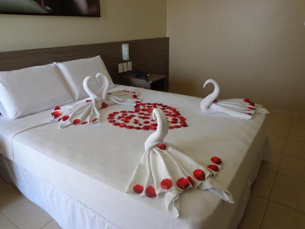 Quarto de hotel com cama bem arrumada, cabeceira de madeira e pequena mesa de cabeceira com abajur.