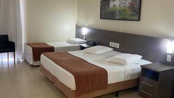Quarto de hotel com duas camas de solteiro, uma mesa de cabeceira com abajur, um pequeno armário e um quadro na parede.
