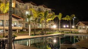 A nighttime view of a hotel pool area with a clear pool, palm trees, and outdoor seating. - Class Hotel Passos Rio Grande Portal da Canastra (Passos)