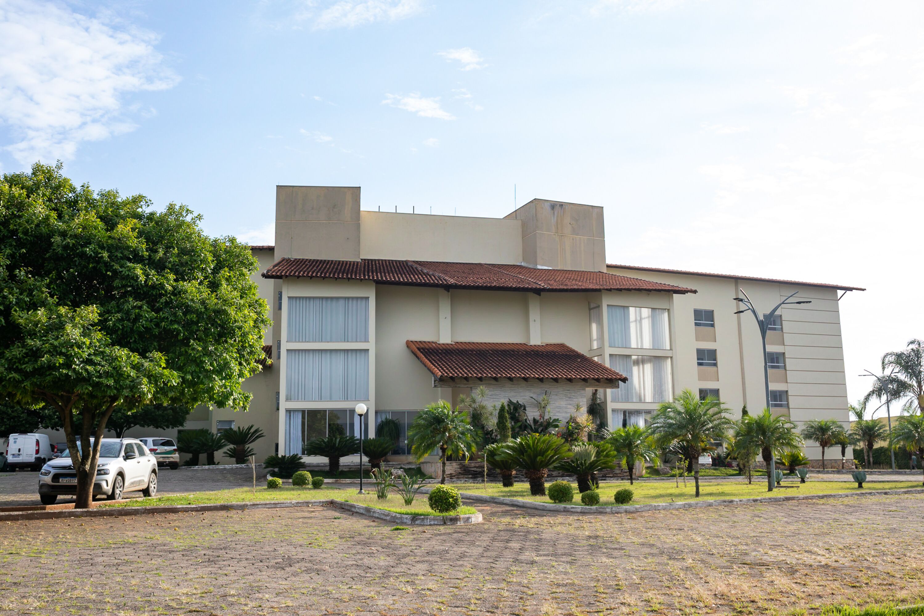 Un hotel de varios pisos con techo plano, ventanas amplias y un patio empedrado con palmeras y arbustos.
