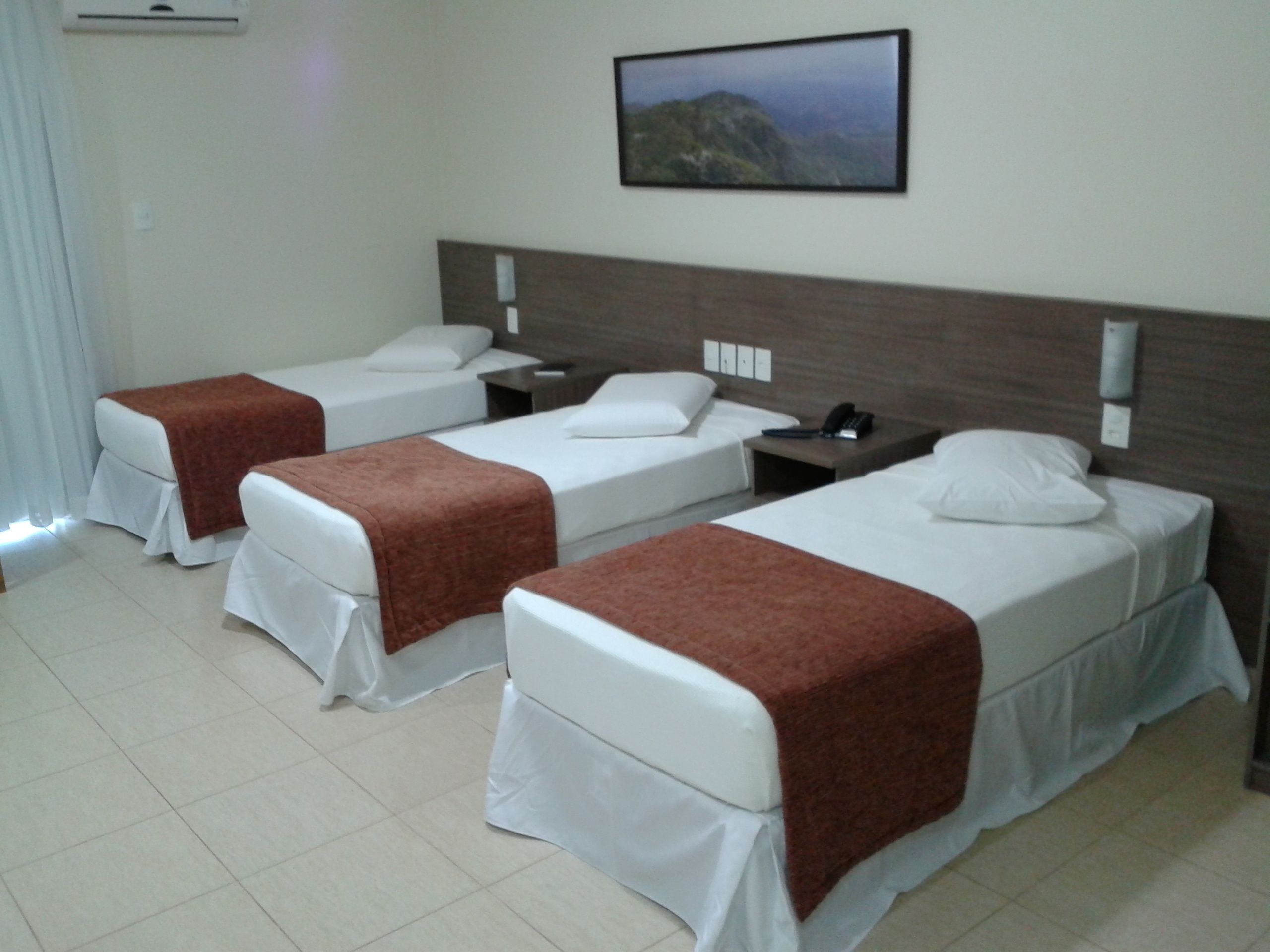 A hotel room with three single beds, each with white bedding and brown headboards. There is a nightstand with a phone and a lamp between the beds. A painting hangs on the wall above the beds.