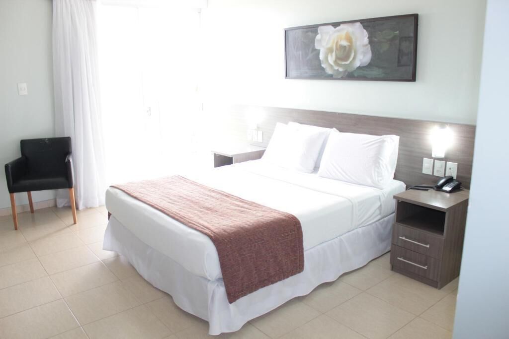 A hotel room with a bed, bedside table, chair, and a painting on the wall.