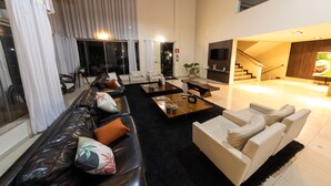 A spacious living room with a black leather sofa, white armchairs, and a coffee table. A staircase leads to an upper level, and a TV is mounted on the wall. - Class Hotel Passos Rio Grande Portal da Canastra (Passos)