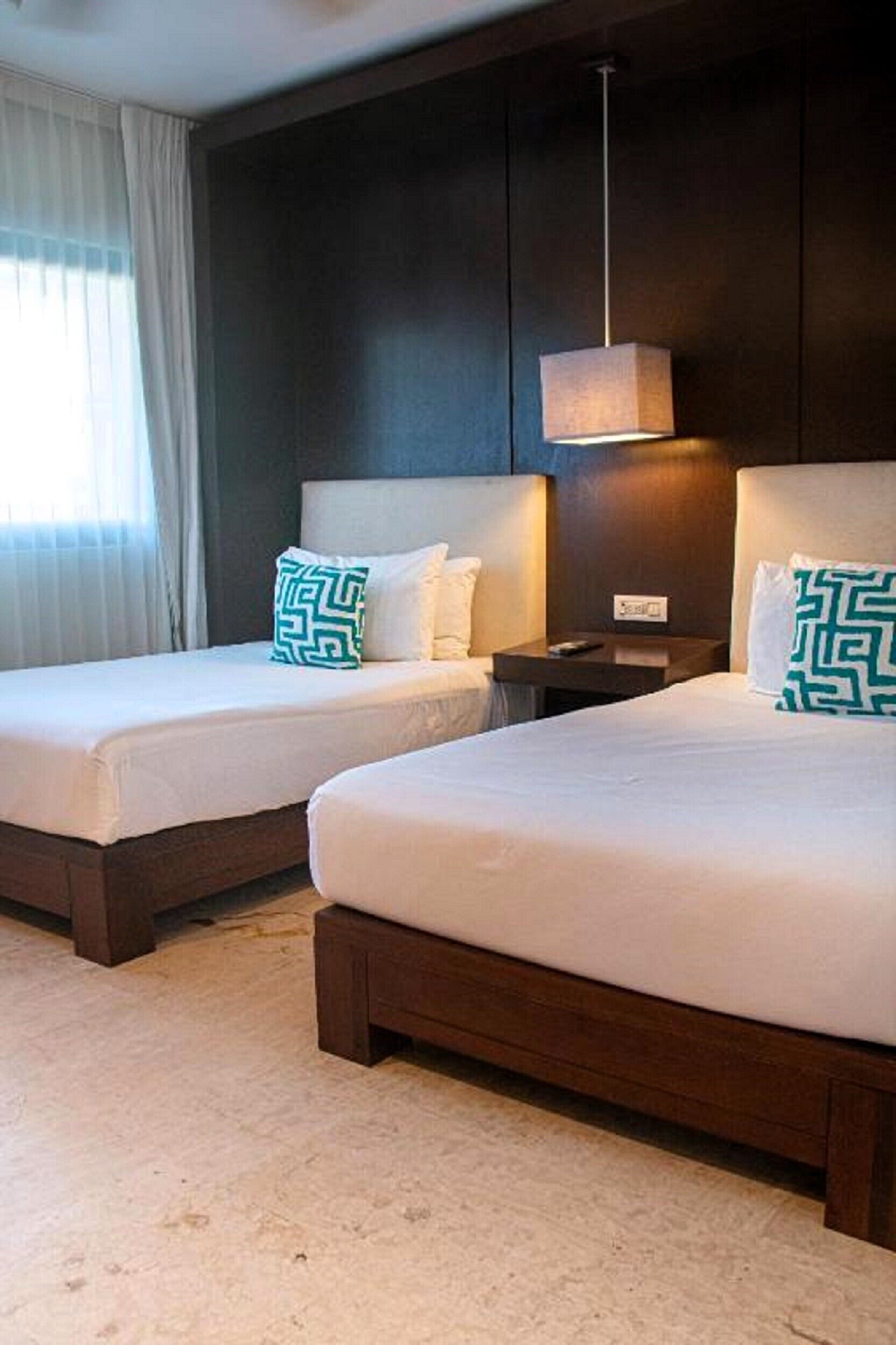 Habitación de hotel con dos camas, una mesita de noche, una lámpara y un ventanal con cortinas.