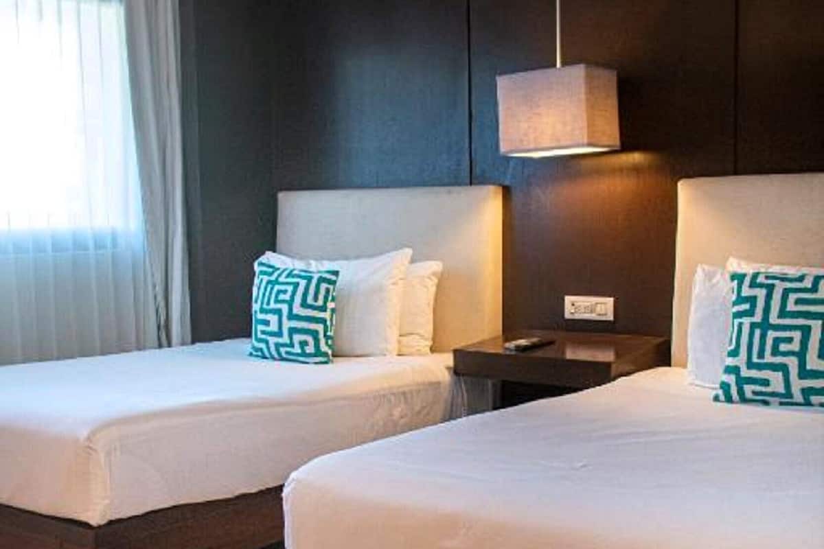 A hotel room with two beds, a nightstand, a lamp, and a window with curtains.