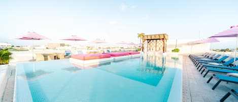 A rooftop pool with clear blue water, sun loungers, and pink-striped umbrellas.