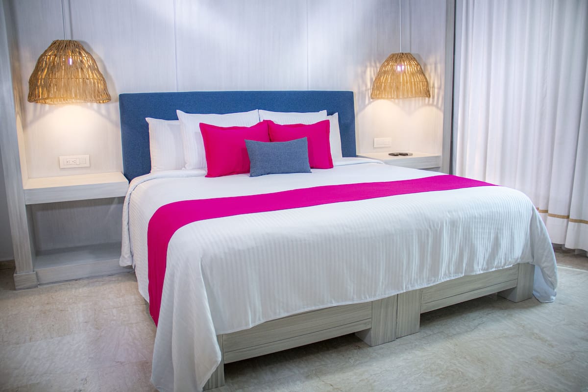 A neatly made bed with a blue headboard, white bedding, and pink and blue pillows. There are two bedside lamps with a woven shade.