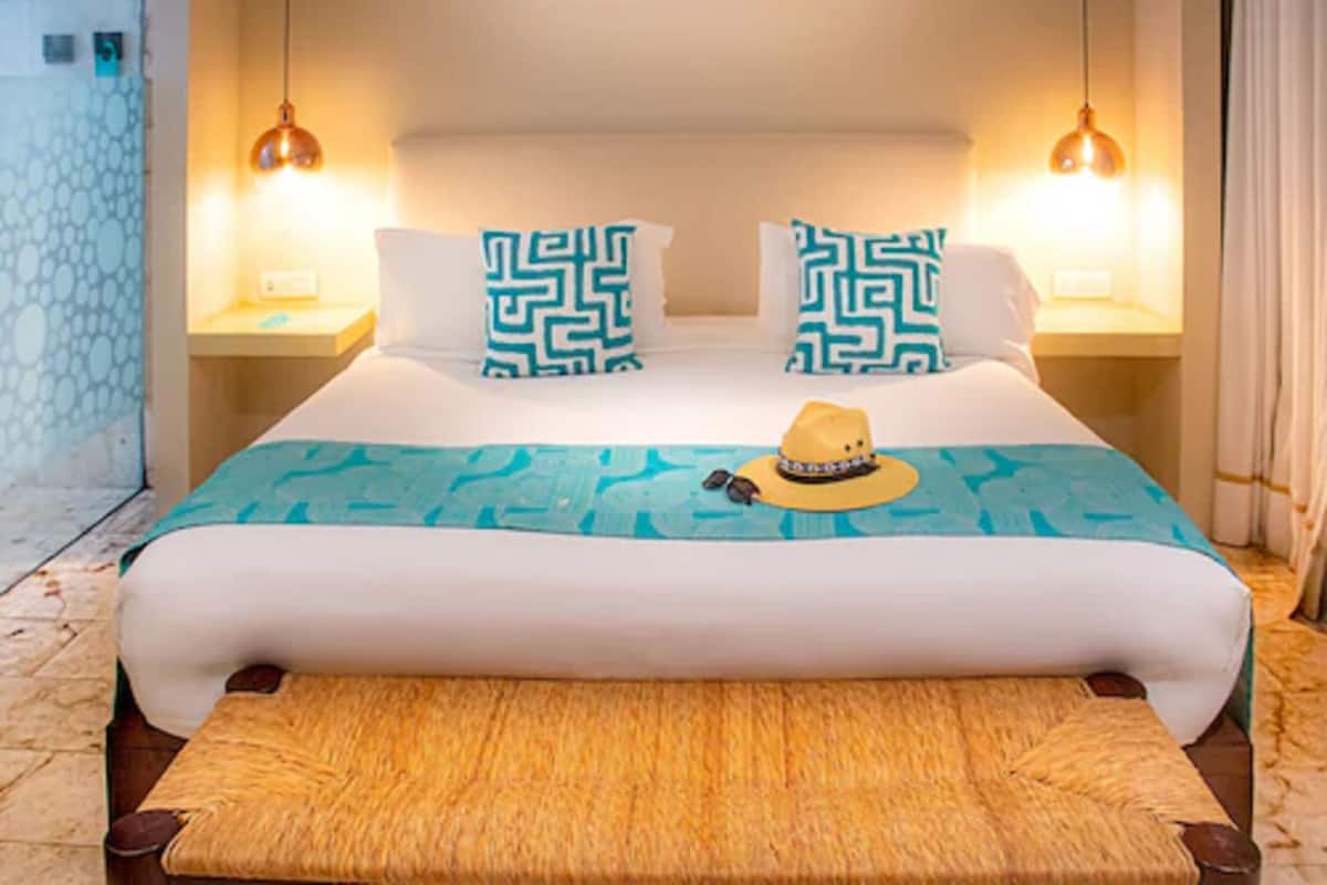 A neatly made bed with patterned pillows, a blue and white bedspread, and a straw hat on the bed. There are two bedside lamps and a nightstand with a lamp on each side of the bed.