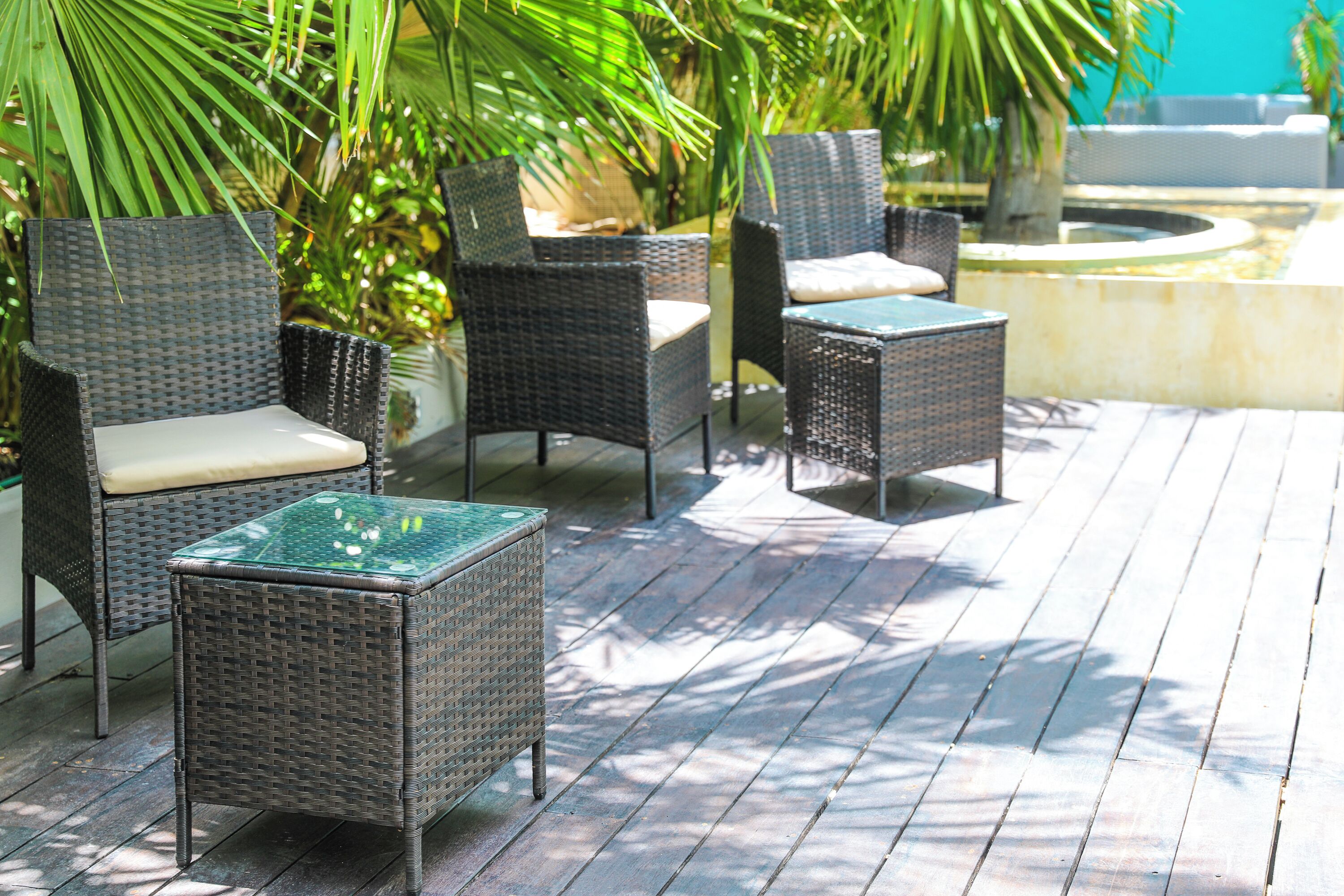 Mobilier d’extérieur en osier, agrémenté de coussins, disposé sur une terrasse en bois, avec une piscine et des palmiers en arrière-plan.
