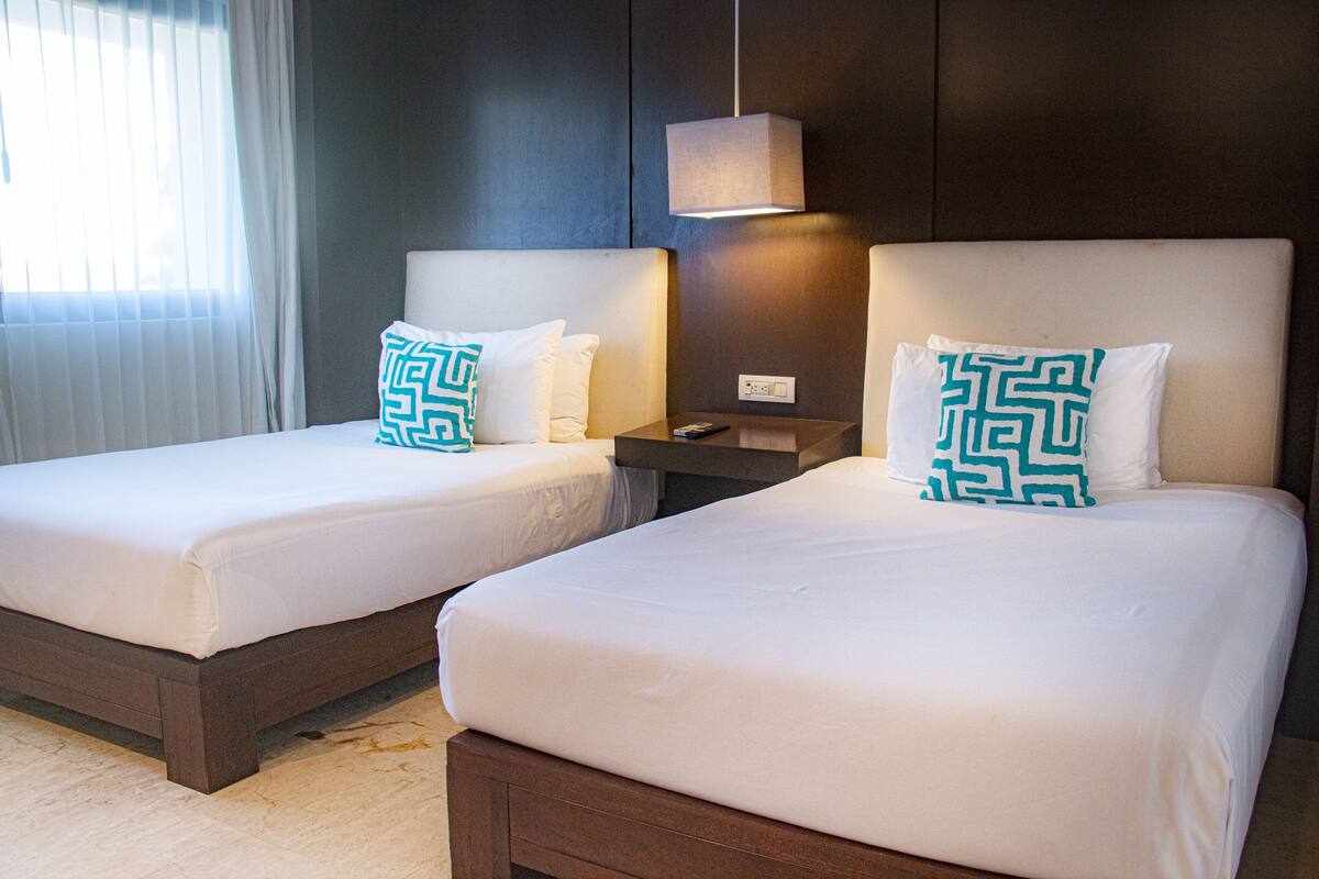 Two twin beds with white bedding and teal patterned pillows, a bedside table with a lamp, and a window with sheer curtains.