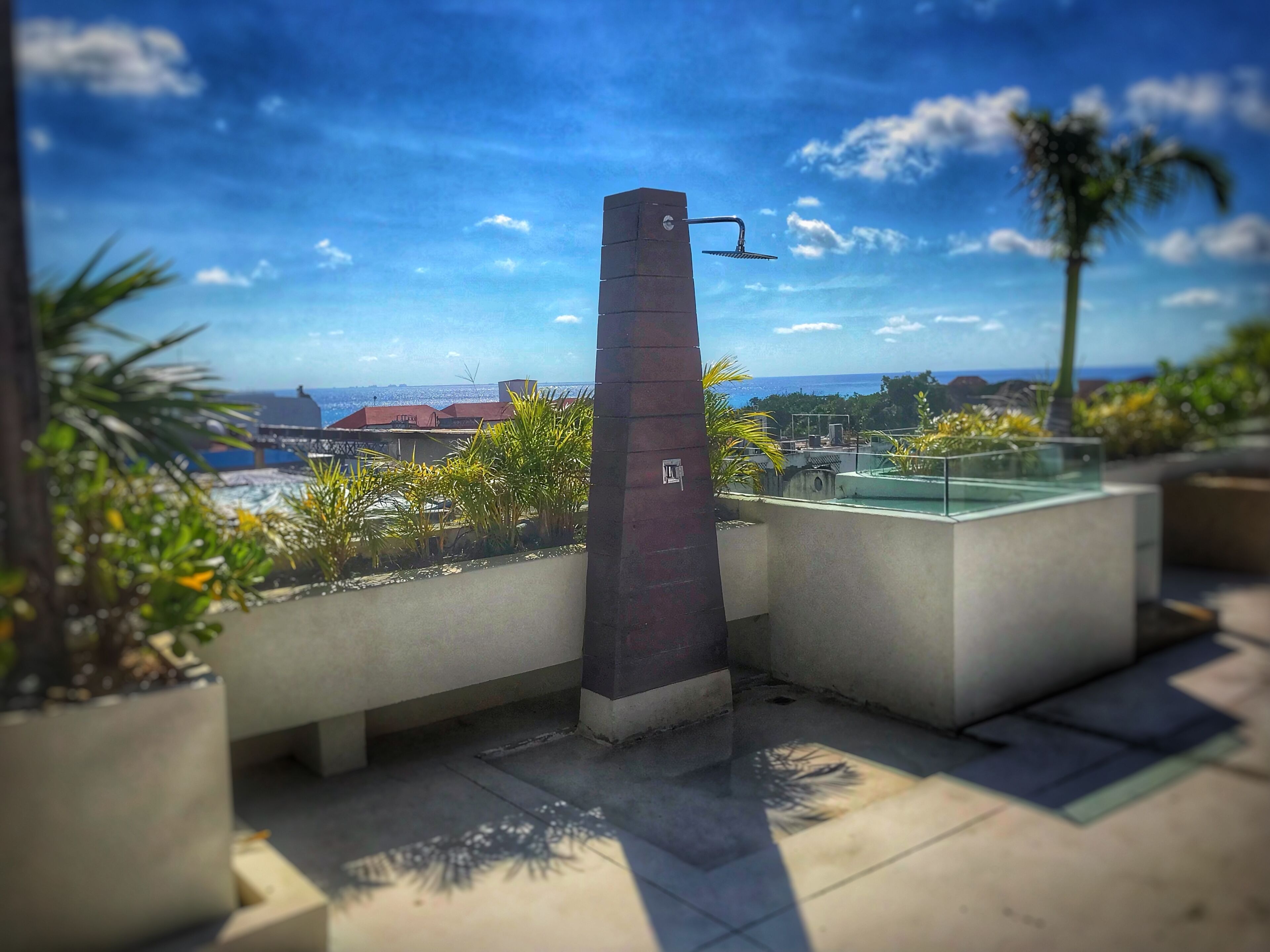 Eine Dachterrasse mit moderner Wasserspielfeatur, Kübelpflanzen und Blick auf das Meer und entfernte Gebäude.