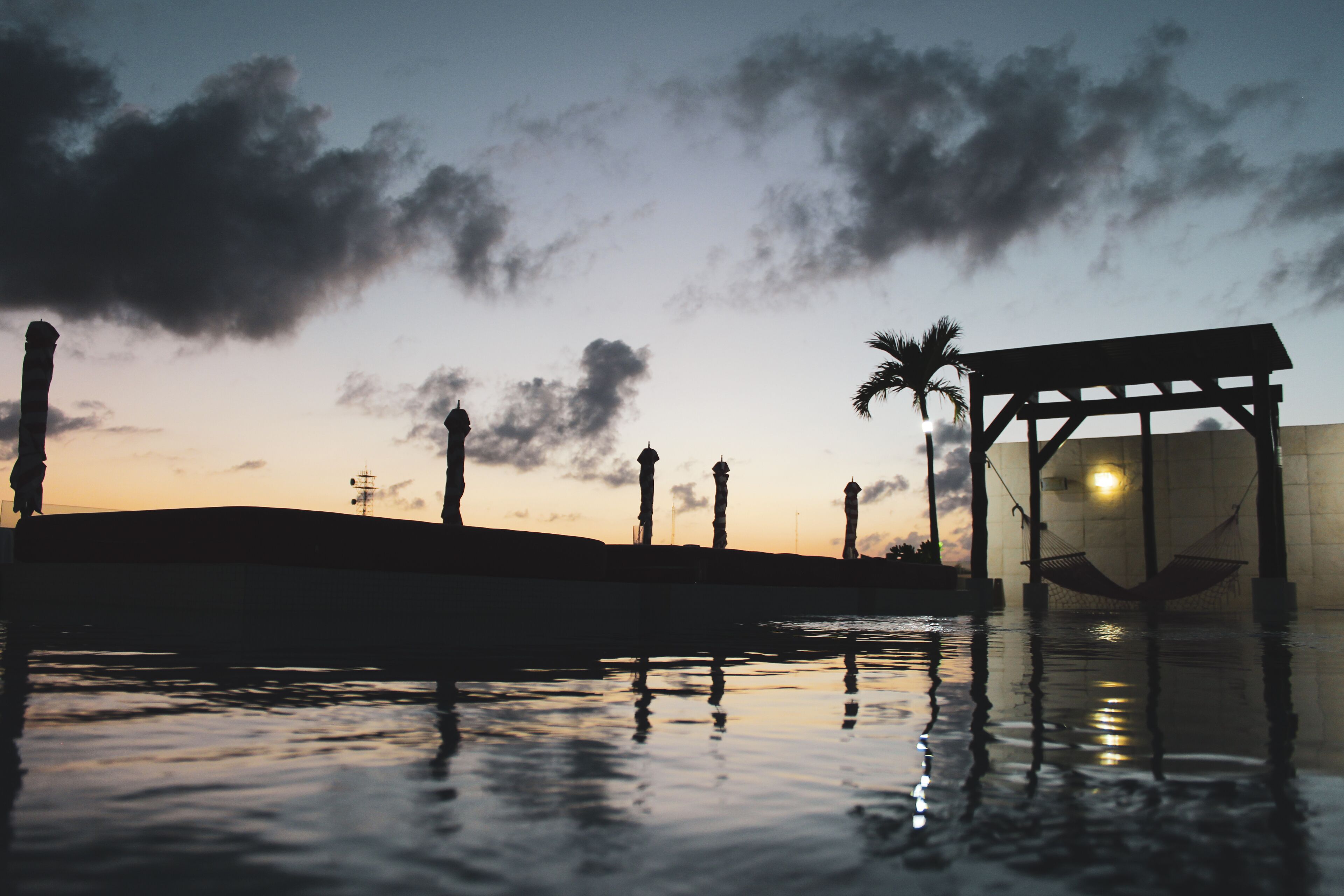 A serene evening by a reflective water body with a gazebo, palm trees, and lamp posts.
