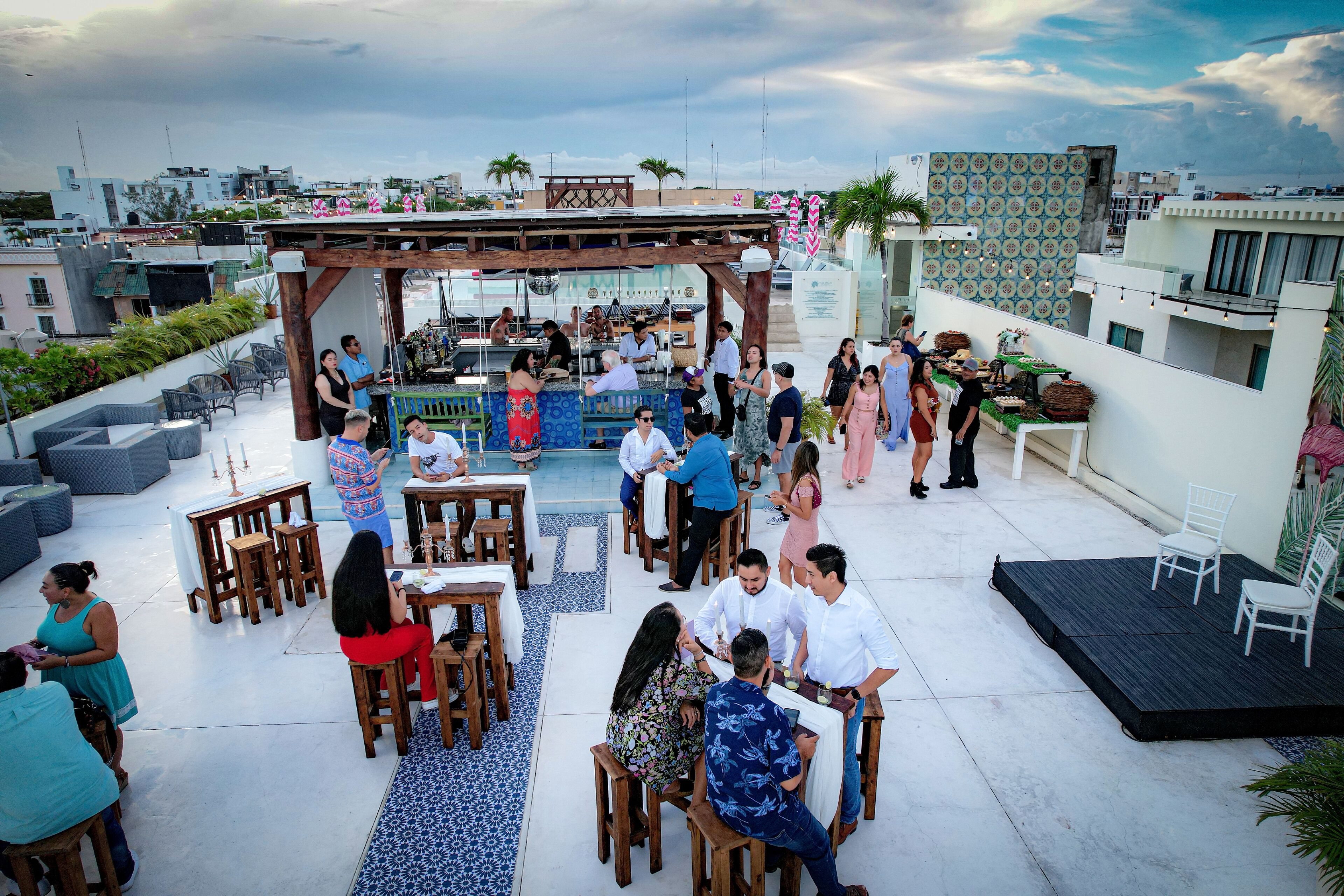 A rooftop bar with wooden tables and chairs, a bar area, and a view of the cityscape.