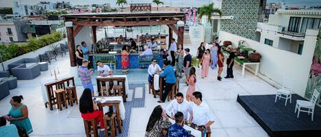 A rooftop bar with wooden tables and chairs, a bar area, and a view of the cityscape.