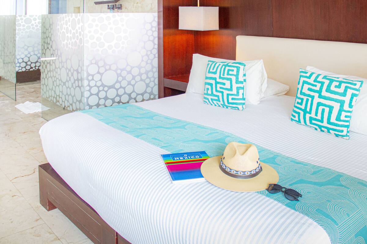 A hotel room with a large bed, patterned pillows, a hat, sunglasses, and a travel guide on the bed.