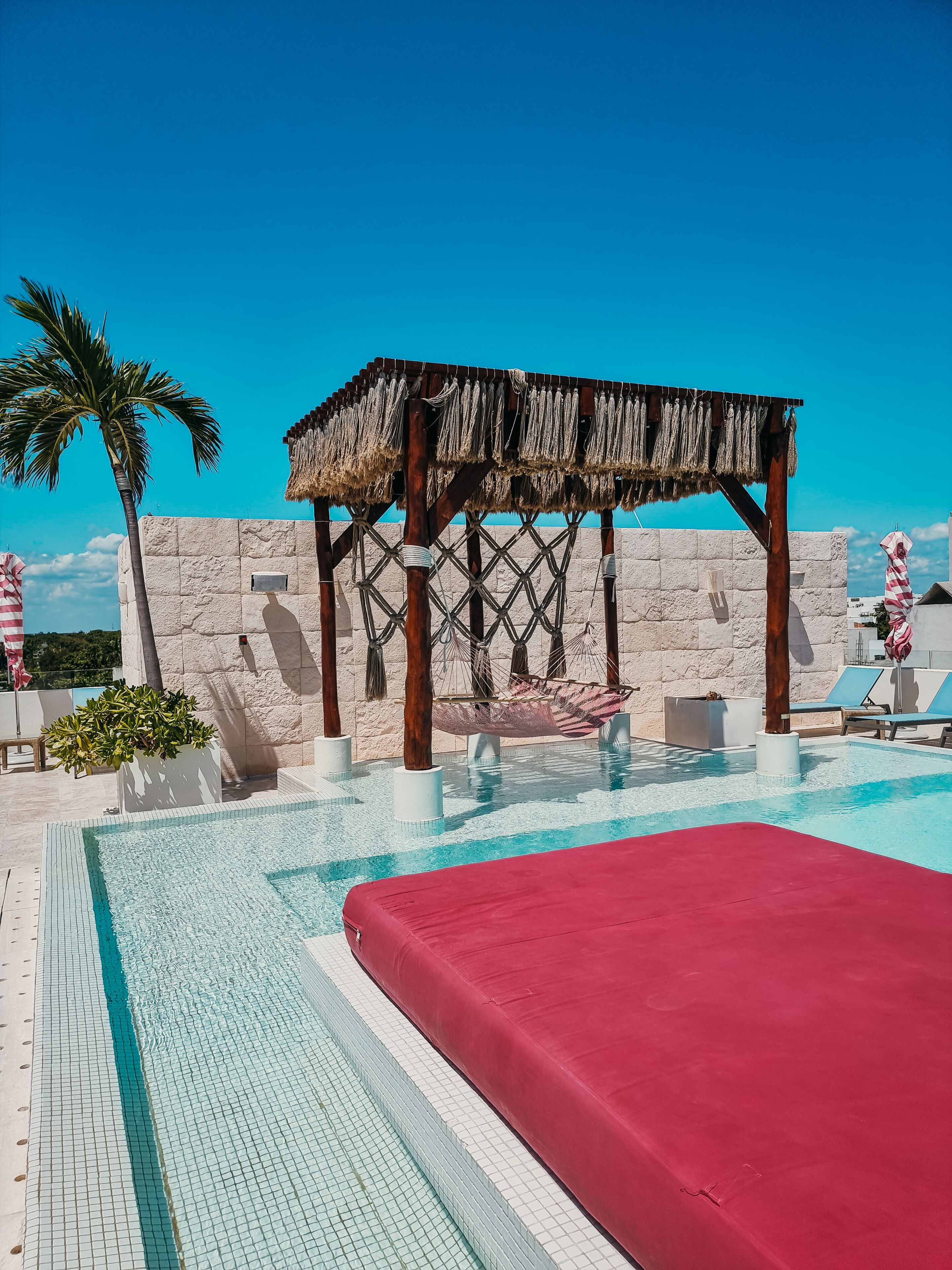 A pool area with a red lounge chair, a thatched structure, and a palm tree.