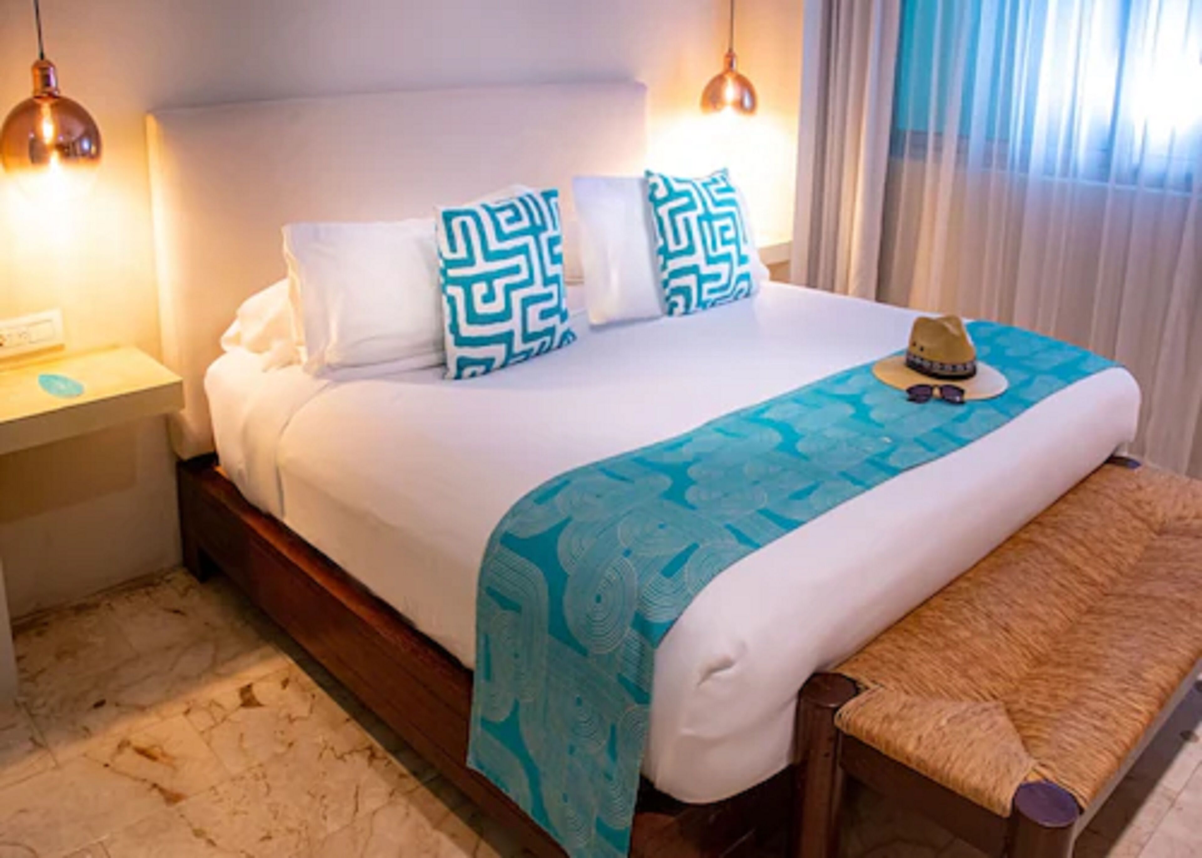 Una cama bien hecha con un edredón estampado, dos almohadas con un diseño geométrico, una lámpara de noche y una mesita con un sombrero y gafas de sol.
