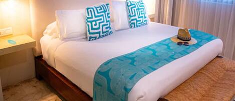 Una cama bien hecha con un edredón estampado, dos almohadas con un diseño geométrico, una lámpara de noche y una mesita con un sombrero y gafas de sol.