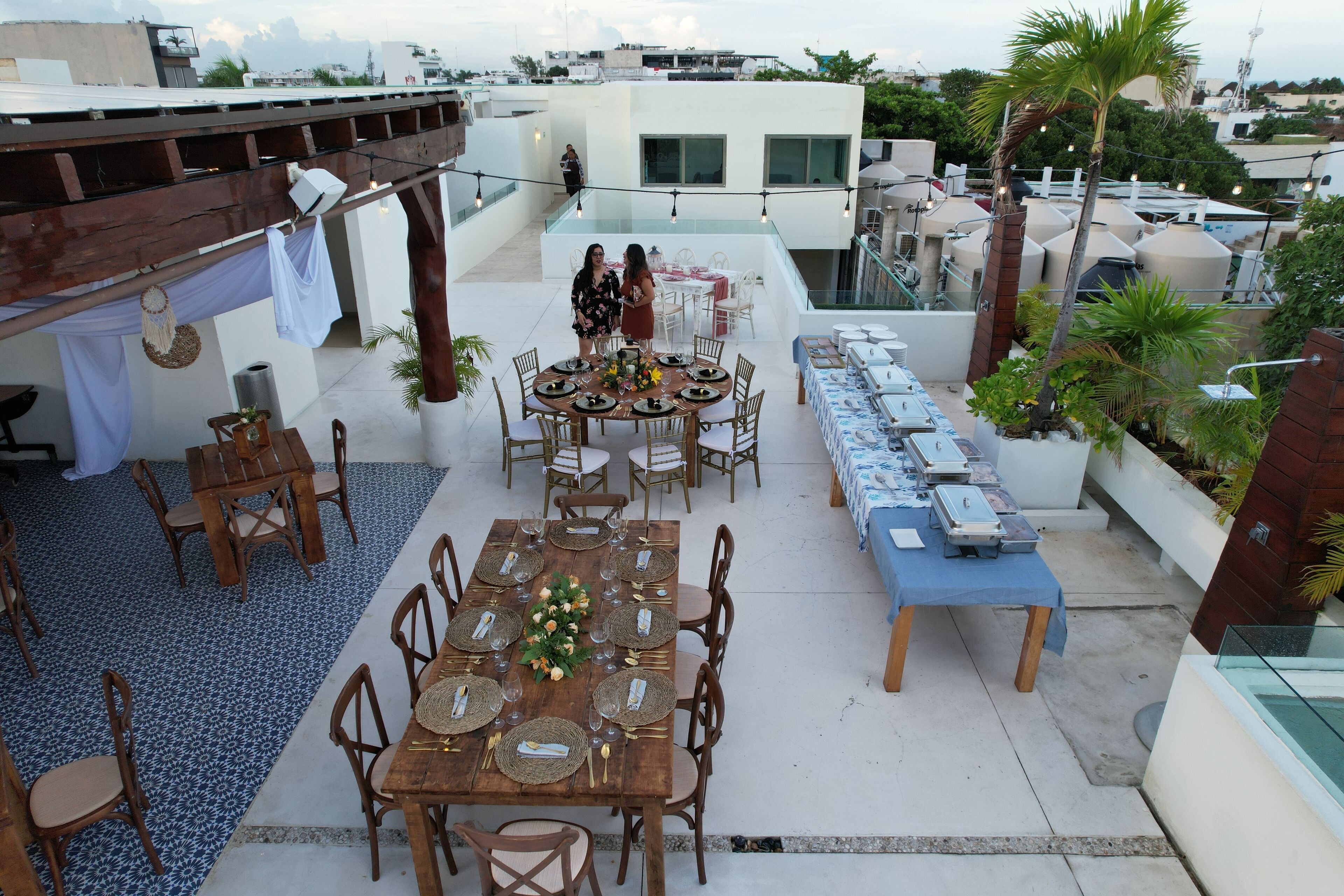 A rooftop terrace with a wooden dining table set for six, surrounded by chairs. There are two people standing near the table. The terrace has a blue patterned floor and is decorated with plants and a string of lights.