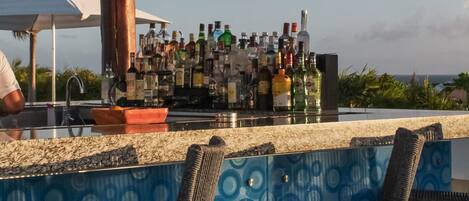 A bar area with a variety of bottles, two wicker chairs, and a view of the ocean.