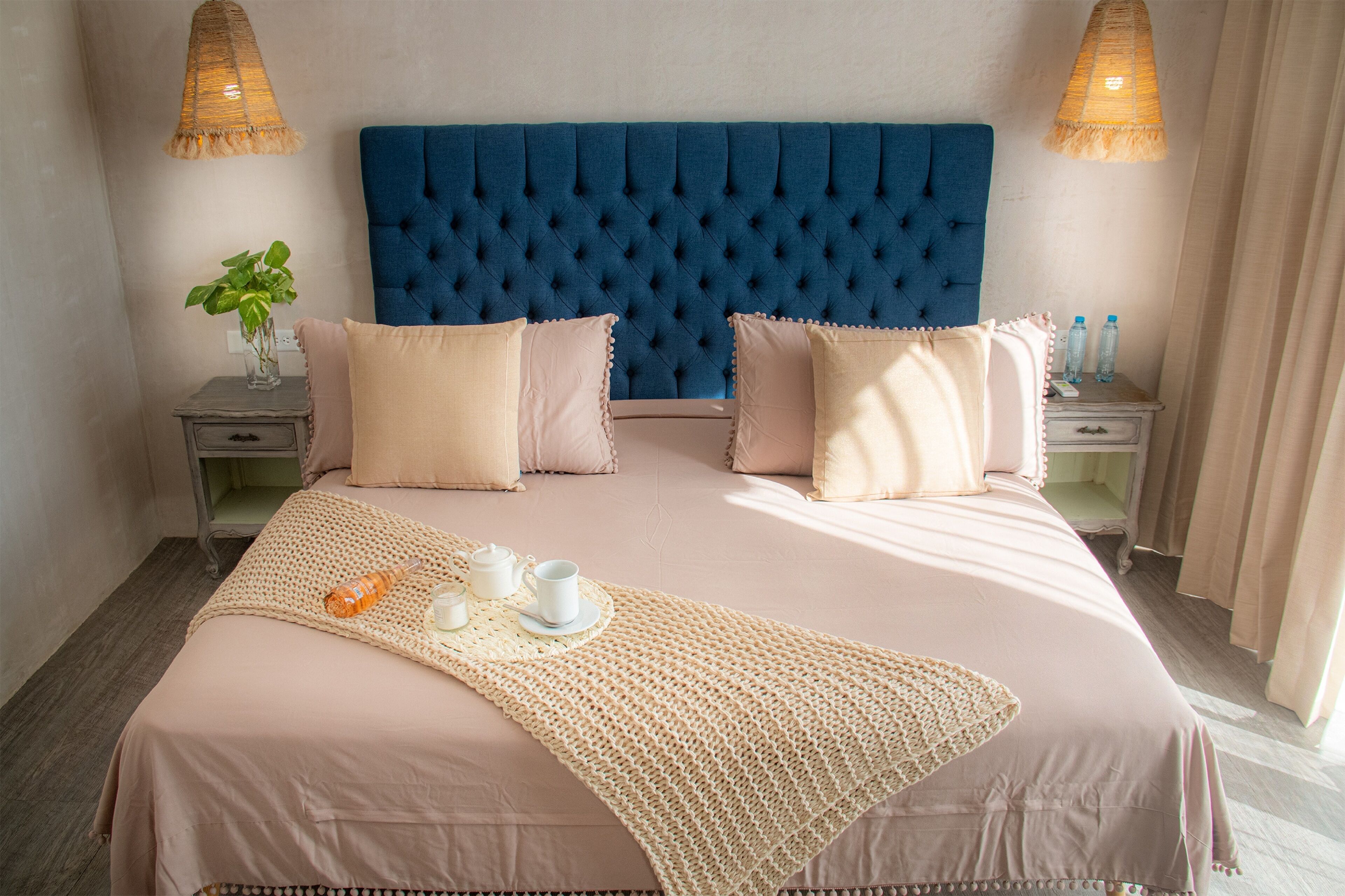 Een net opgemaakt bed met een blauw hoofdbord, twee nachtkastjes met lampen en een geweven plaids.