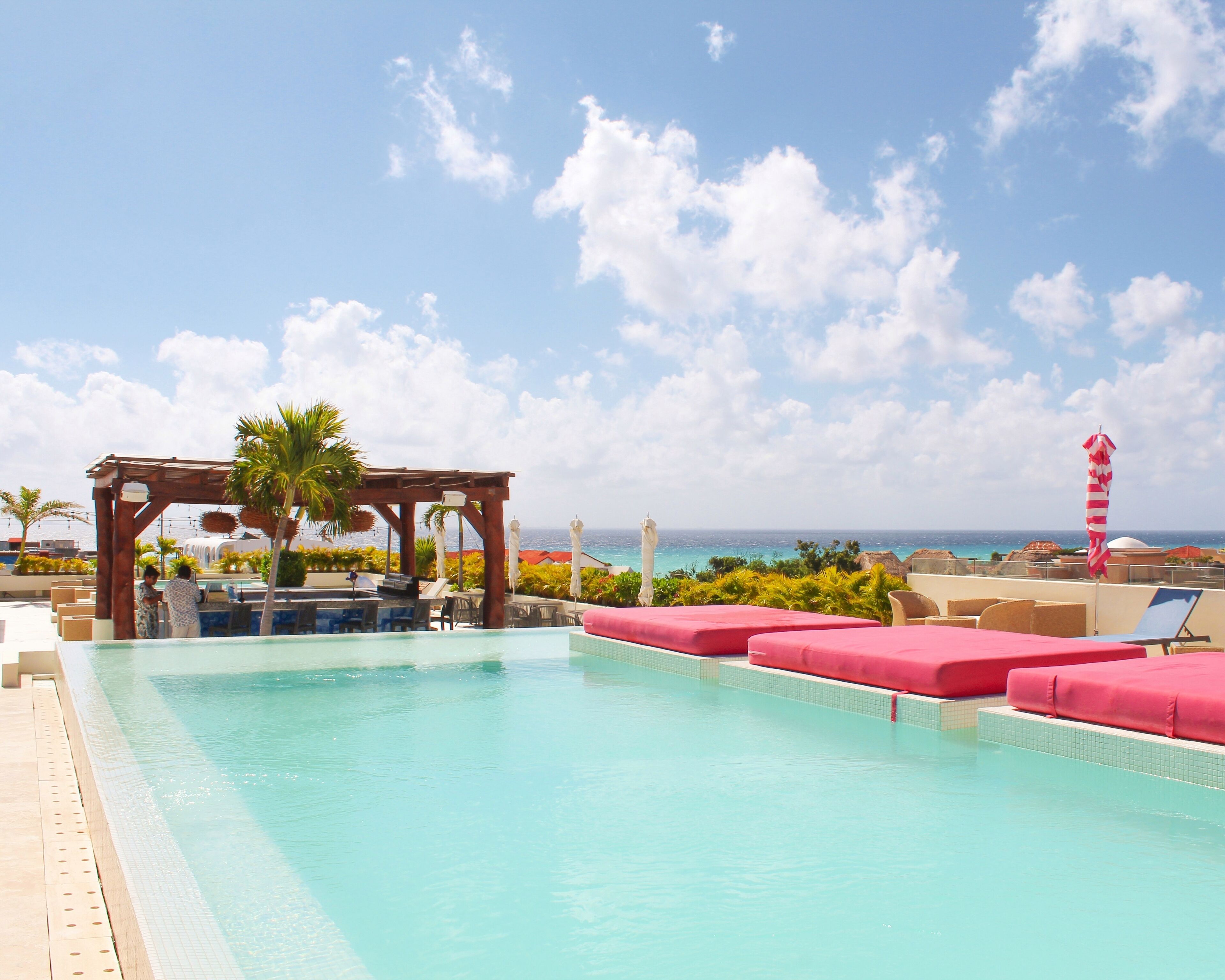 A pool area with lounge chairs and a wooden pergola, overlooking a beach and ocean.