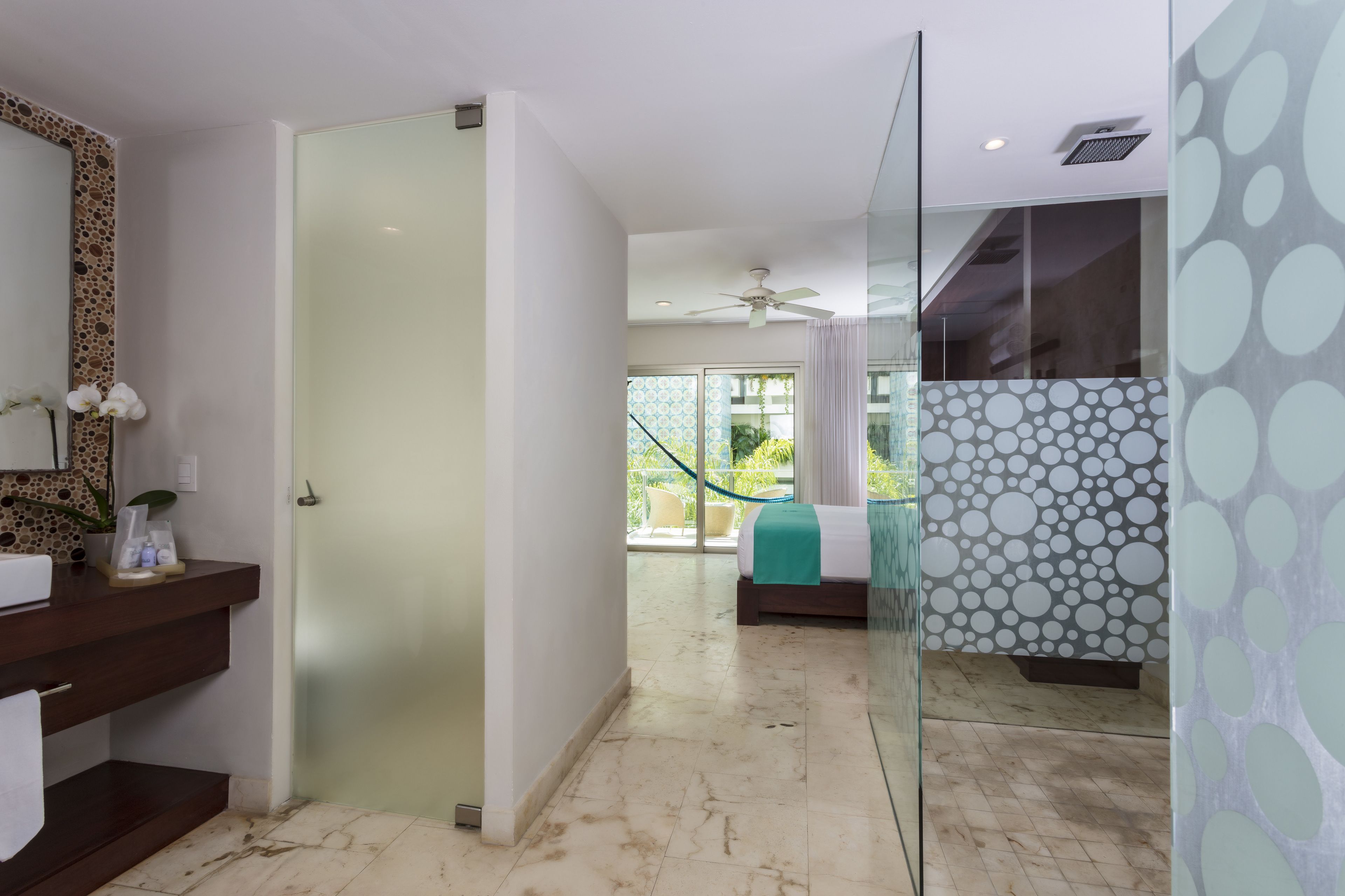A modern hotel room with a glass shower, a bed with a green bedspread, a bathroom with a marble floor, and a view of the outdoors.