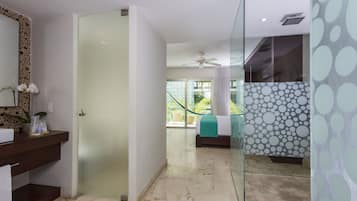 Quarto de hotel moderno com chuveiro de vidro, cama com colcha verde e banheiro com piso de mármore, além de vista para o exterior.