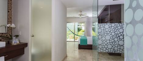 A modern hotel room with a glass shower, a bed with a green bedspread, a bathroom with a marble floor, and a view of the outdoors.