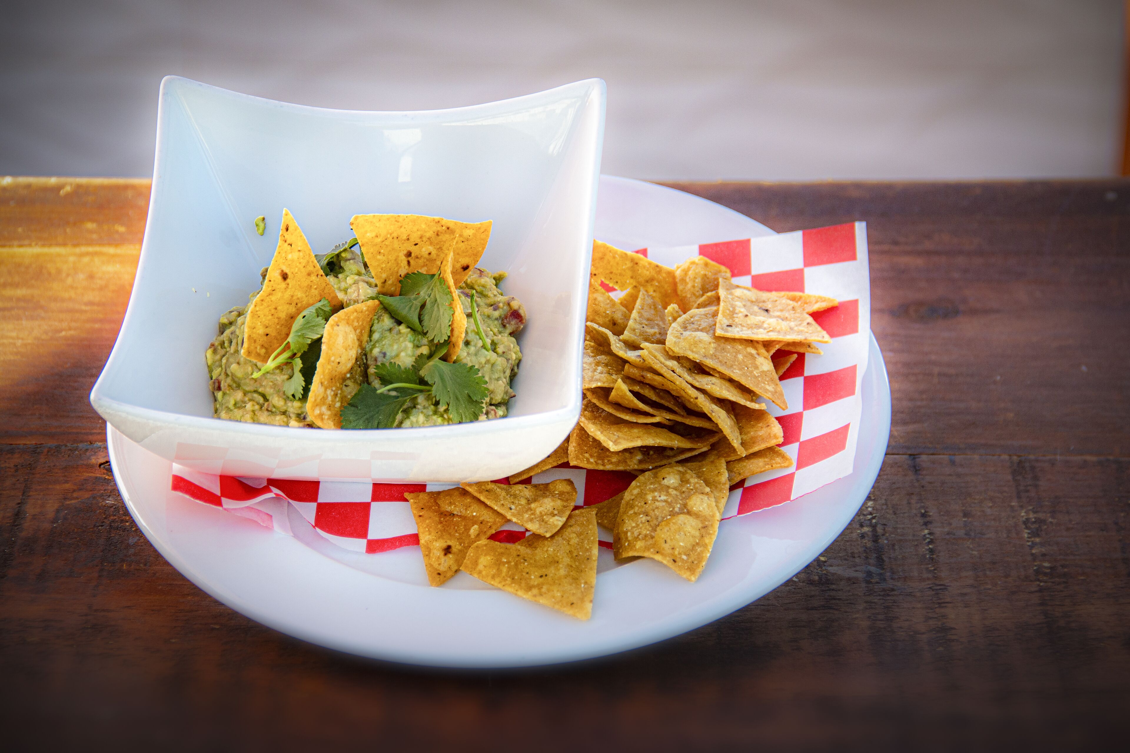 A white bowl of guacamole with tortilla chips on a white plate with red and white checkered paper.