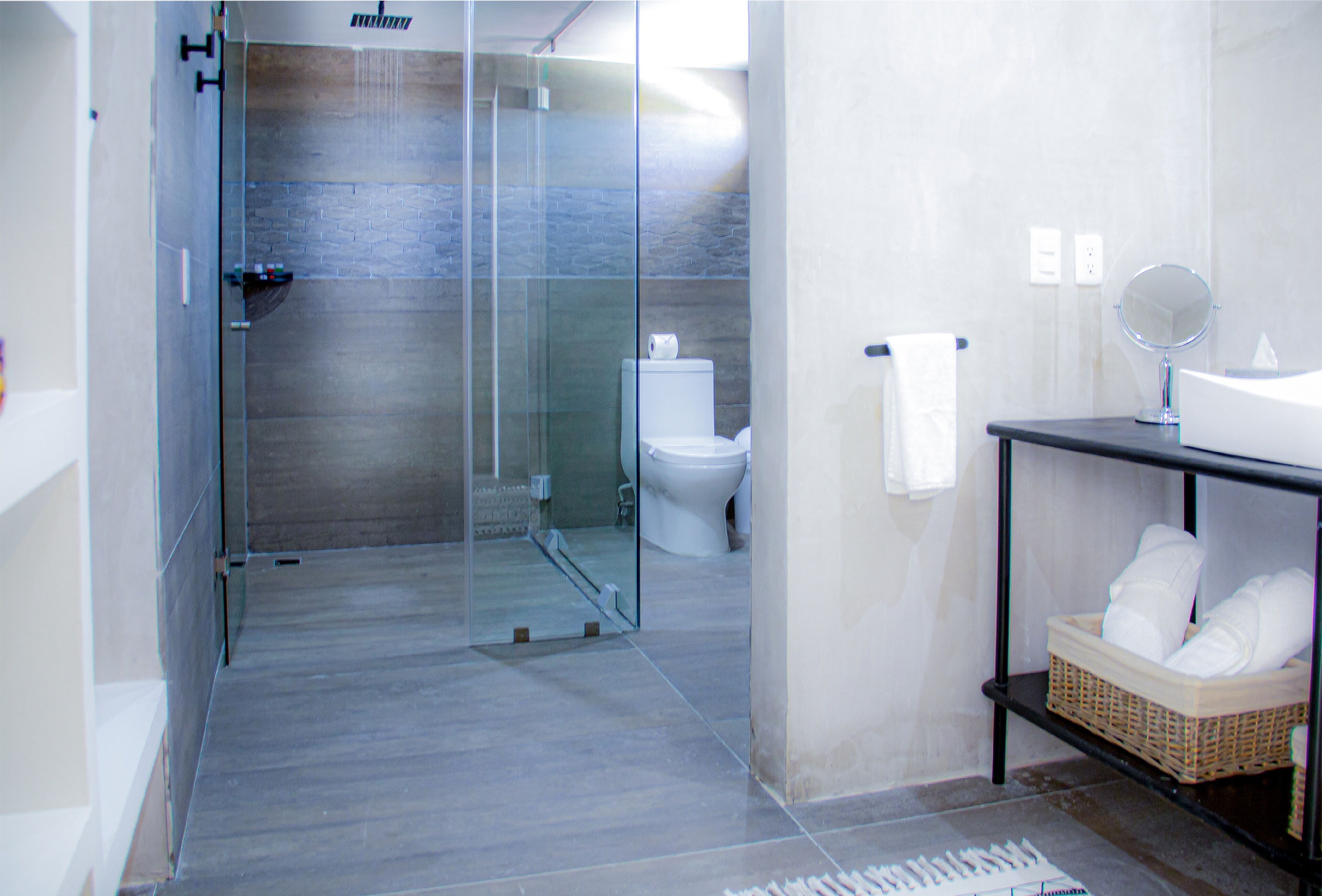 A modern bathroom with a glass shower enclosure, a white toilet, a sink with a mirror, and a basket of towels on a shelf.