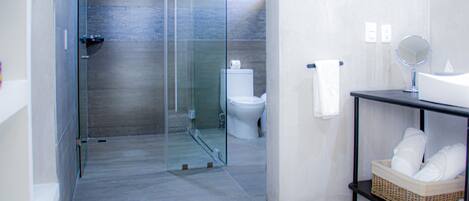 A modern bathroom with a glass shower enclosure, a white toilet, a sink with a mirror, and a basket of towels on a shelf.