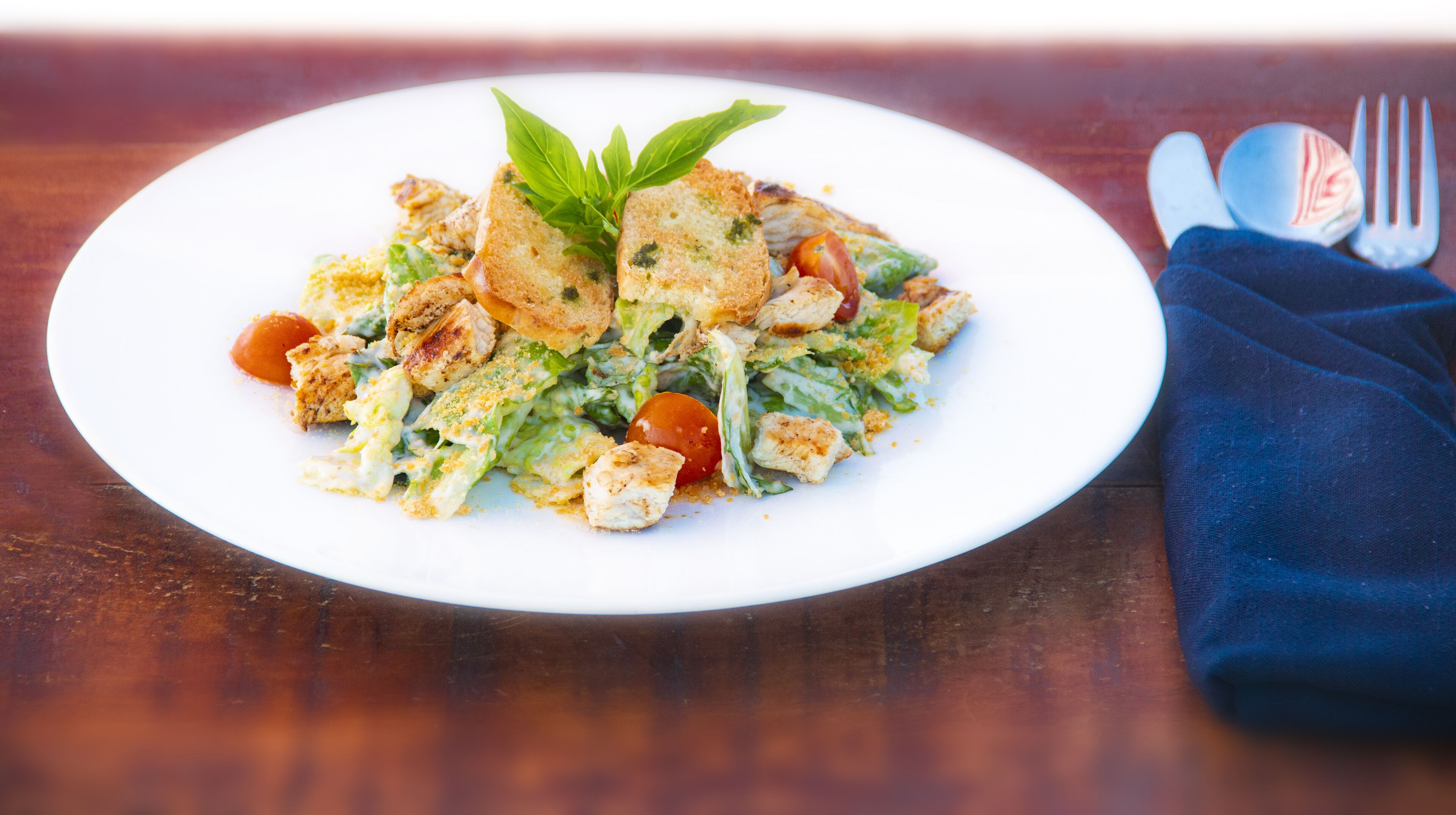 A white plate with a salad, croutons, and a basil leaf on a wooden table.
