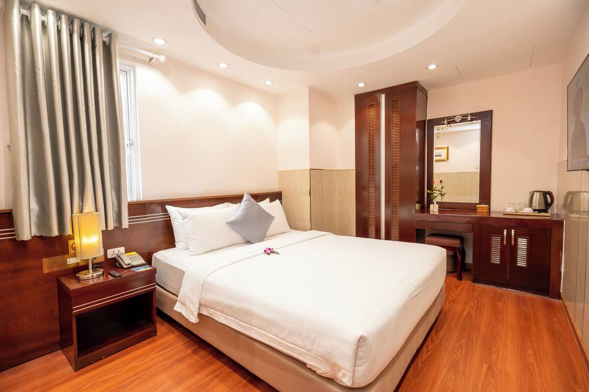 A hotel room with a large bed, a bedside table with a lamp, a desk with a mirror, and a wardrobe.