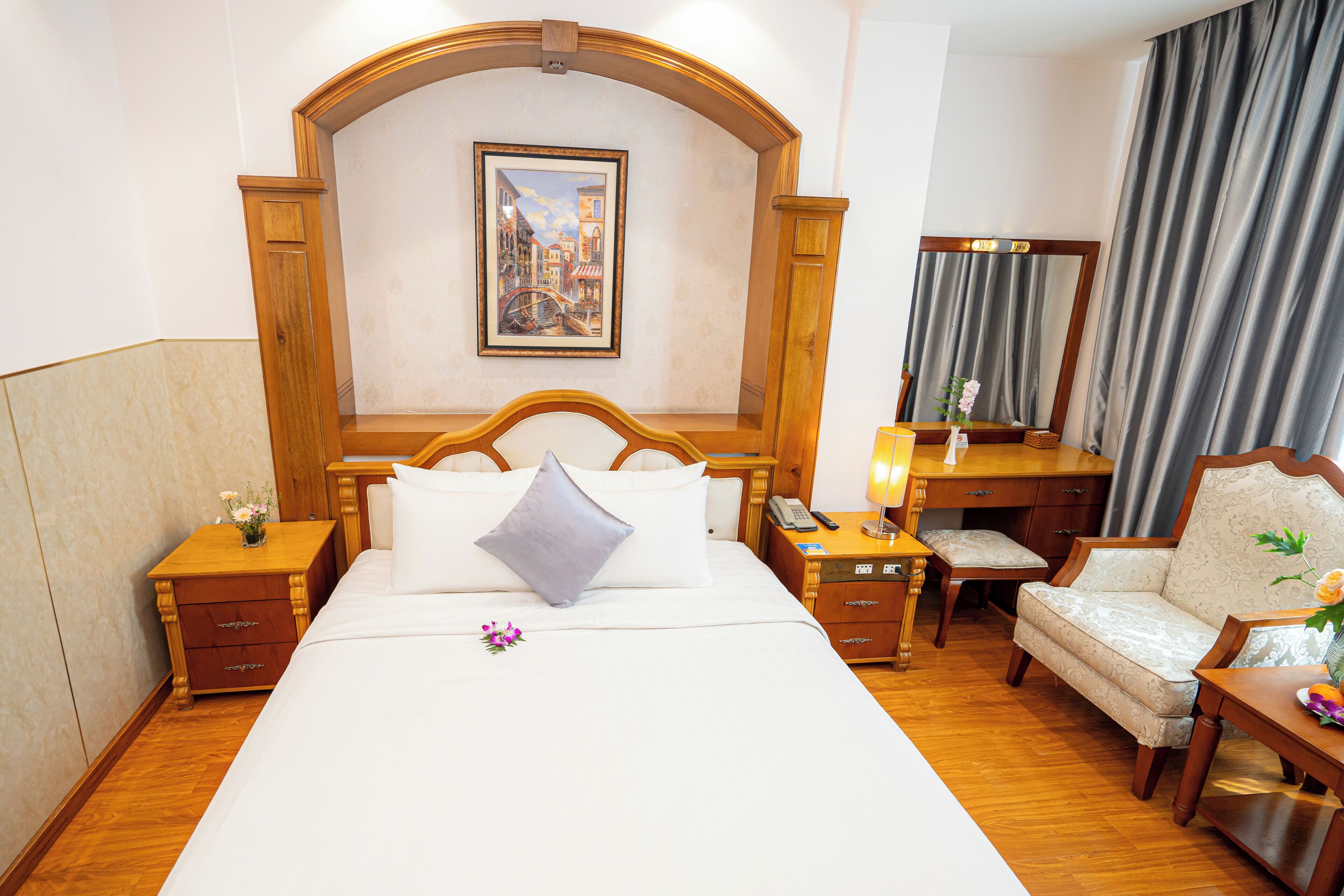 A hotel room with a large bed, a wooden headboard, a bedside table with a lamp, a chair, a desk with a mirror, and a painting on the wall.