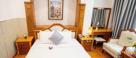 A hotel room with a large bed, a wooden headboard, a bedside table with a lamp, a chair, a desk with a mirror, and a painting on the wall.