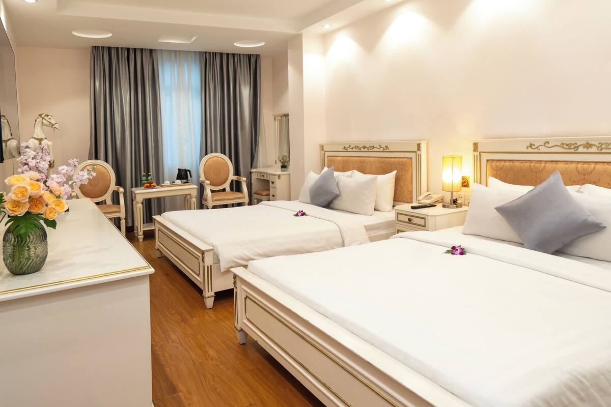 A hotel room with two beds, a desk, chairs, a vanity, and a large window with curtains.