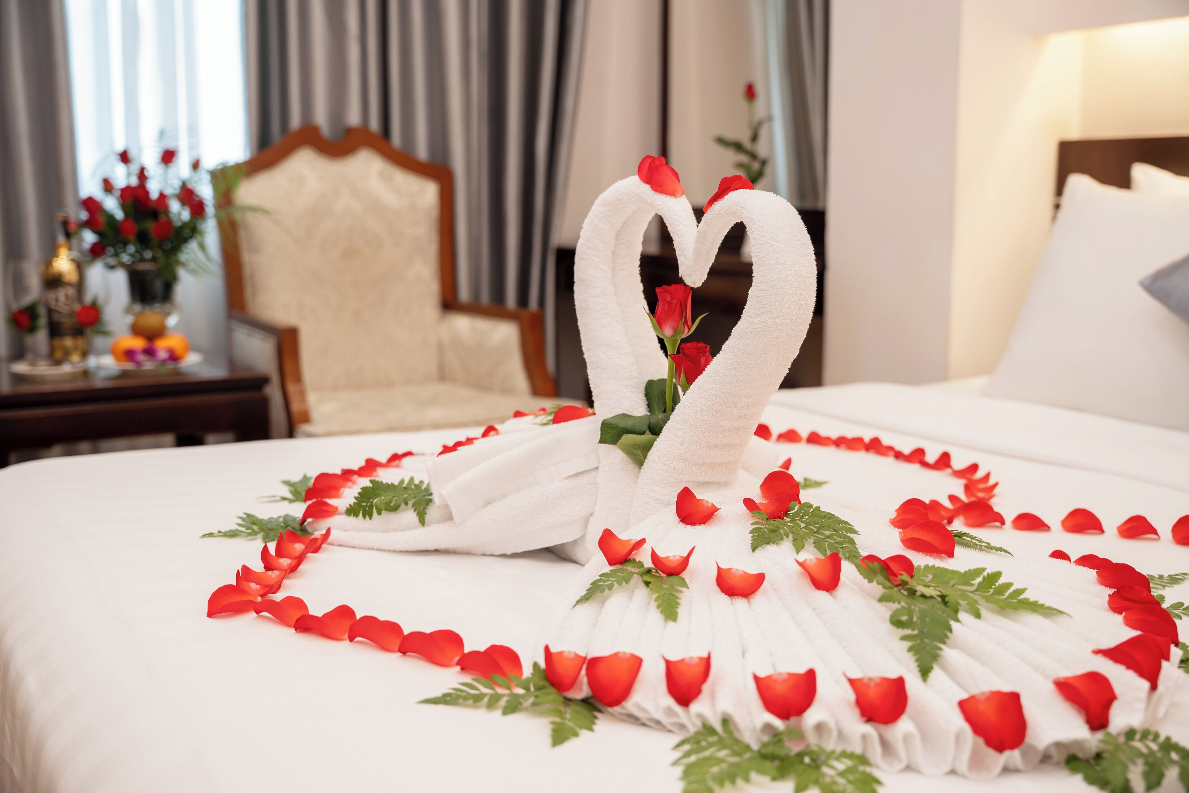 A hotel room with a bed adorned with a heart-shaped towel arrangement and rose petals.