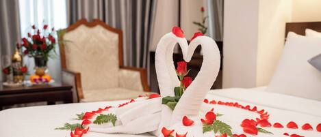 A hotel room with a bed adorned with a heart-shaped towel arrangement and rose petals.