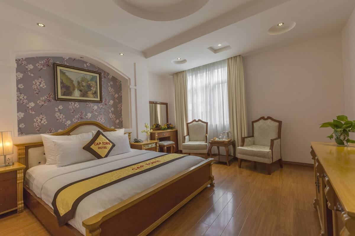 A hotel room with a large bed, two armchairs, a wooden desk, a mirror, and a floral wallpaper.