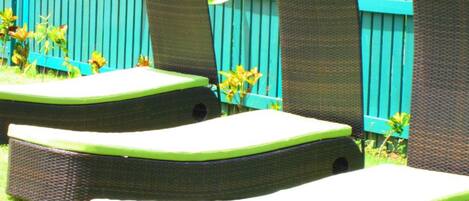Three outdoor lounge chairs with green cushions, arranged in a row on a grassy lawn, with a blue picket fence in the background.