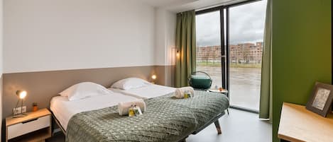 A hotel room with two beds, a desk, a chair, a window with a view of buildings, and a green wall.
