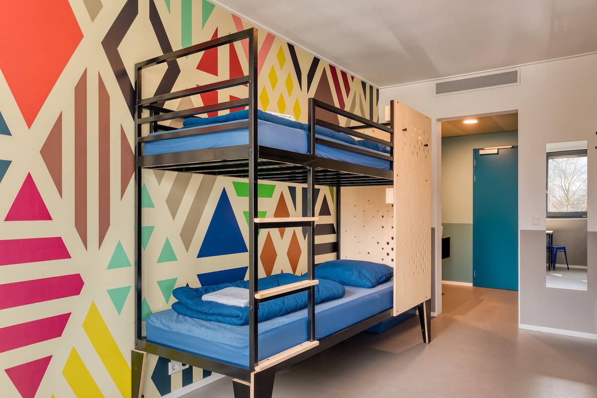 Une chambre avec des lits superposés, un mur orné d’un motif géométrique, une porte en bois et une vue sur un couloir.