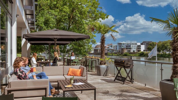 An outdoor patio area with seating, a grill, and a view of the water. - Stayokay Maastricht - Hostel (Maastricht)