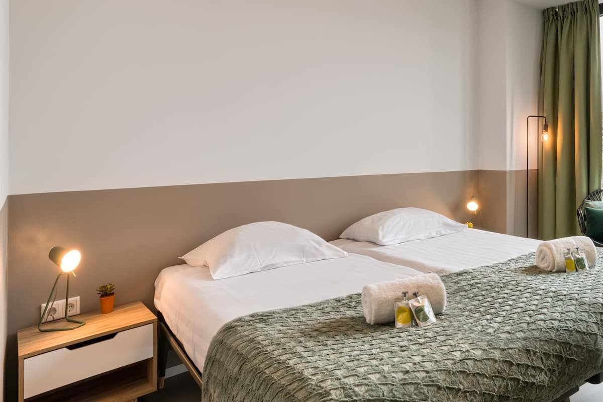 Un lit bien fait, avec du linge de lit blanc, une couverture verte à texture intéressante et deux serviettes enroulées accompagnées de produits de toilette. On aperçoit une table de chevet avec une lampe et une petite plante.