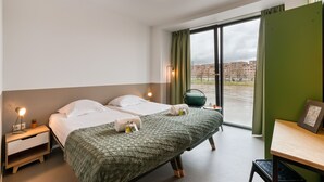 A hotel room with a bed, bedside table, lamp, and a view of the city through the window. - Stayokay Maastricht - Hostel (Maastricht)